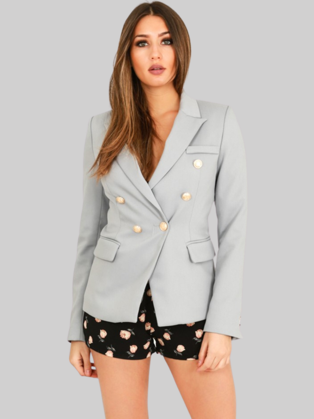 female model wears a grey double breatsted blazer with gold military buttons at the front of the blazer and the cuffs. The model has tanned skin and long brown hair, the model faces the camera and wears the military button blazer closed. The model has both hands to her sides and looks directly into the camera. 
