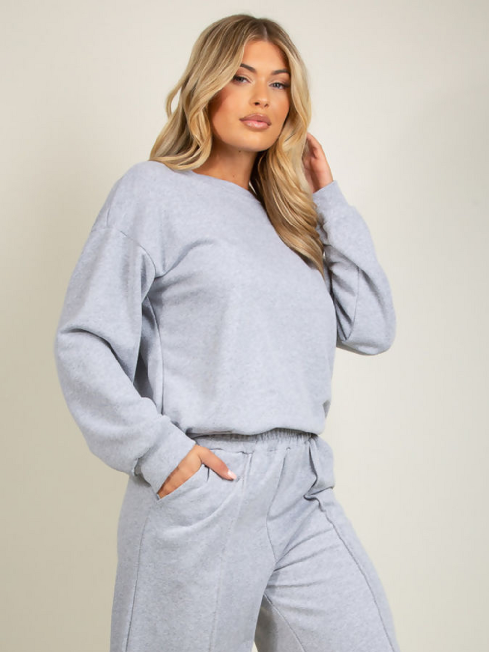 Model wears grey round neck long sleeve jumper and elasticated wide leg matching joggers. The joggers has a pockets anddecorative seam. Model has blonde hair and stares into the camera and stands slightly to the side. Model places one hand in her pocket and the other to her head. 