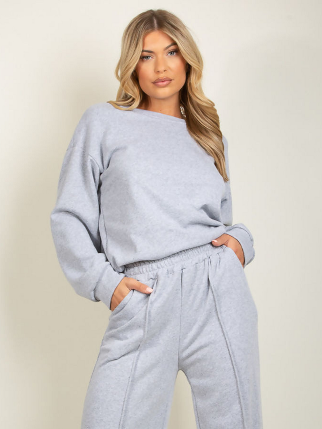 Model wears grey round neck long sleeve jumper and elasticated wide leg matching joggers. The joggers has a pockets anddecorative seam. Model has blonde hair and stares directly into the camera. 