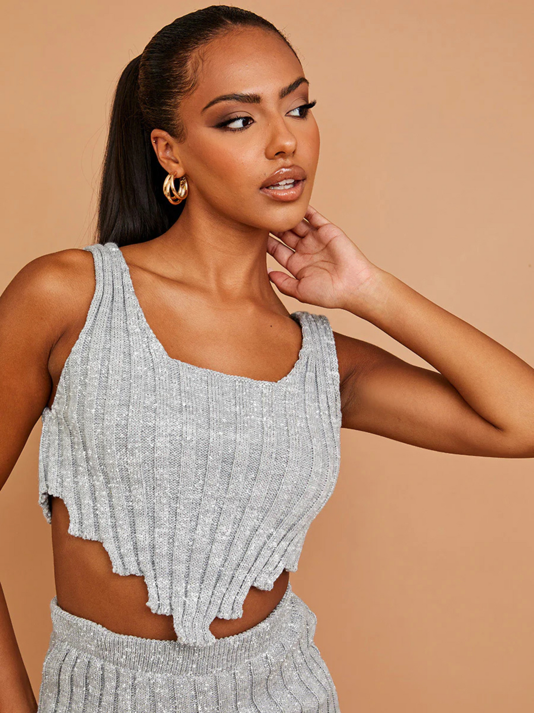 Model wears a grey knitted crop top with a jagged asymmetric hem and square  neckline. The top has a a silver flexedlurex detailing woven into the garment. The Top has an elasticated matching skirt. The model has brown skin and black hair that is pulled back into a long ponytail, she stands looking off to the side, the crop top and top of the matching skirt can be seen. 