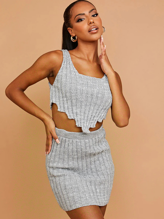 Model wears a grey knitted crop top with a jagged asymmetric hem and square  neckline. The top has a a silver flexedlurex detailing woven into the garment. The Top has an elasticated matching skirt. The model has brown skin and black hair that is pulled back into a long ponytail, she faces the camera, the front of the top and  the atching skirt can be seen. The model laces one hand on her hip and touches her chin with her other hand. 