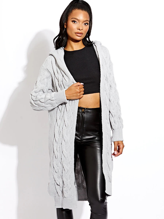 Female model stands in front of a plain background. The female model wears a long open cable knit Cardin in grey. The grey cable knit cardigan has long sleeves. The model has light brown skin and long black hair, she stands with one hand on the cable knit cardigan and the other by her side. 