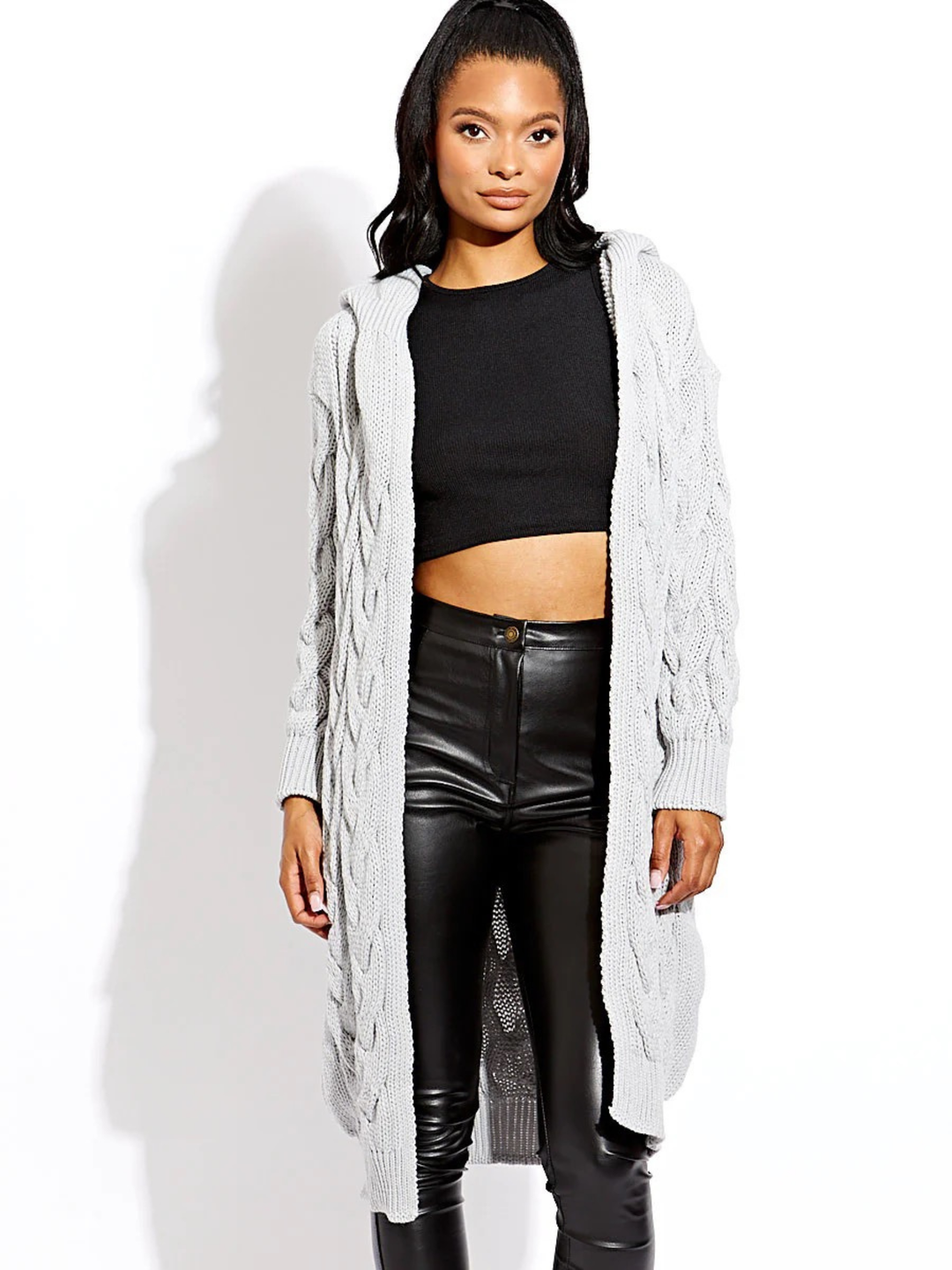 Female model stands in front of a plain background. The female model wears a long open cable knit Cardin in grey. The grey cable knit cardigan has long sleeves. The model has light brown skin and long black hair, she stands with both hands by her sides and looks directly into the camera. 