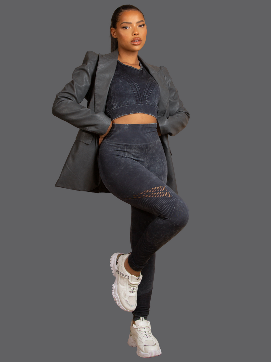 Full length shot. Model wears of Grey Faux Leather Long Sleeved Blazer, With Front Pocket Look and Faux Leather Lapel. Model has light brown skin and a long pony tail. Model faces the camera, she wears the grey leather look blazer and active wear. 