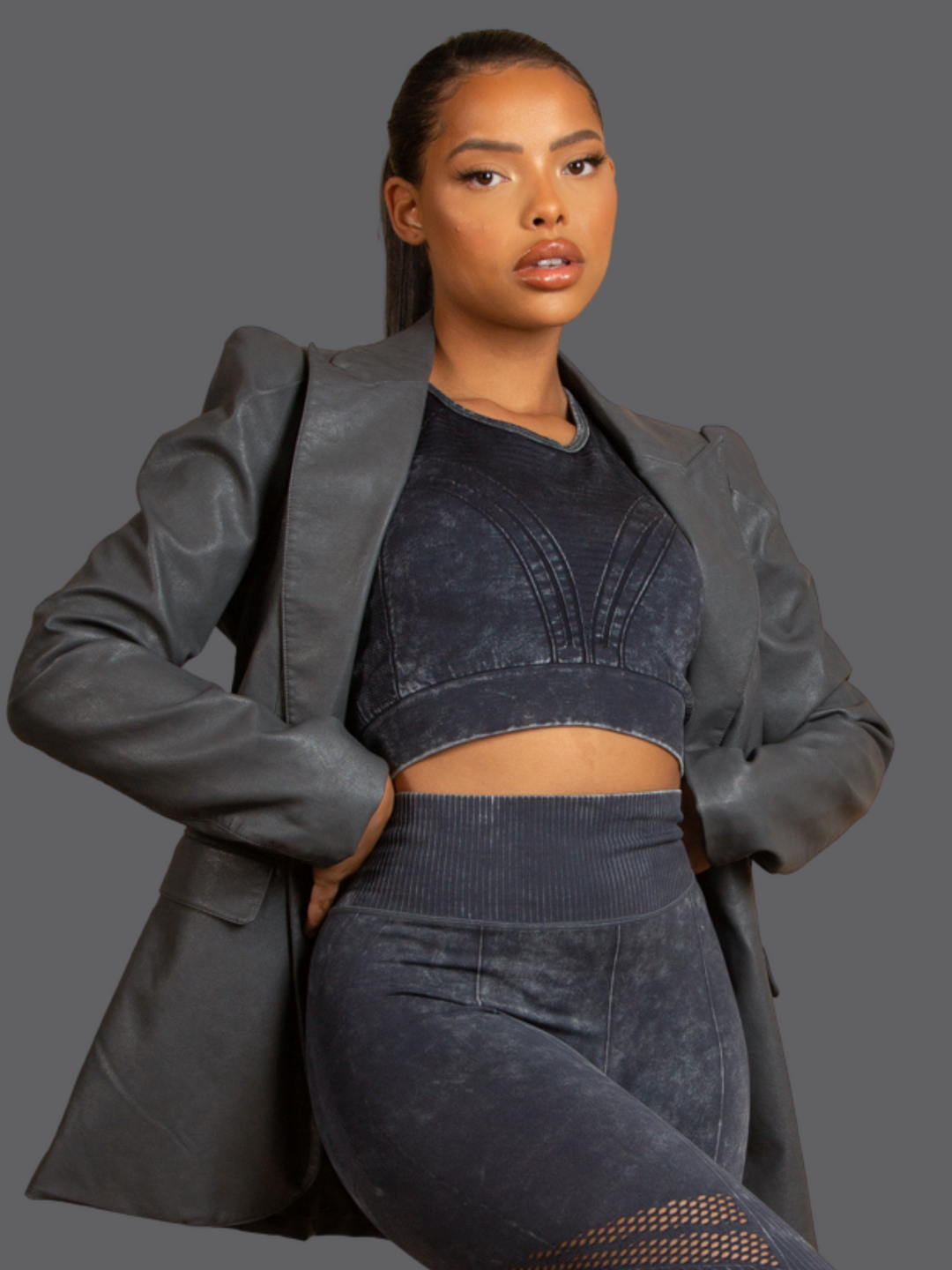 Model wears of Grey Faux Leather Long Sleeved Blazer, With Front Pocket Look and Faux Leather Lapel. Model has light brown skin and a long pony tail. Model places her hands on either side of her waist. 