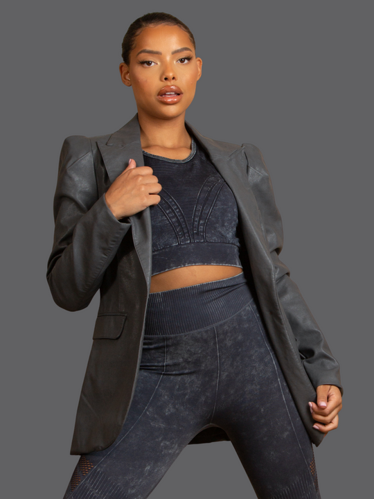 Model wears of Grey Faux Leather Long Sleeved Blazer, With Front Pocket Look and Faux Leather Lapel. Model has light brown skin and a long pony tail. Model stands with one hand to the side and holds the faux leather lapel with the other hand. 