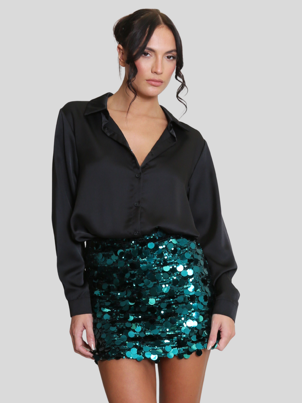 Model wears a luxe disc sequin mini skirt in green and a black shirt. The green  sequin mini skirt,  has a zip closure fastening. The model has tanned skin and black hair, she places her hands by her sides