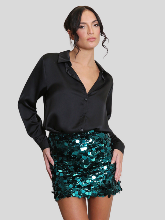 Model wears a luxe disc sequin mini skirt in green and a black shirt. The green  sequin mini skirt,  has a zip closure fastening. The model has tanned skin and black hair, she faces the camera and places her arms by her sides, the luxe sequin skirt reflects the light.
