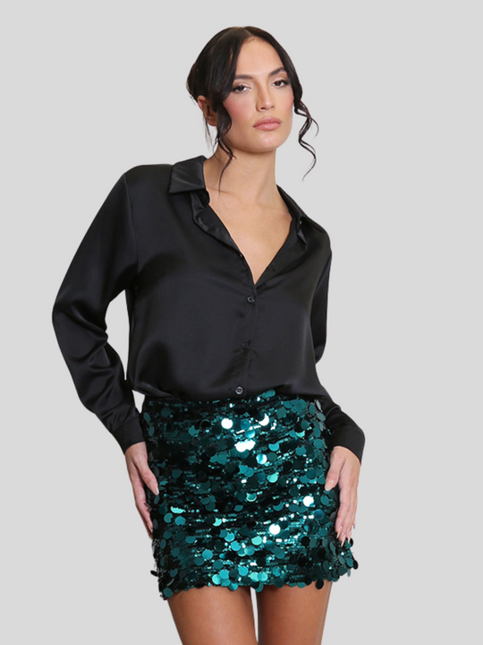 Model wears a luxe disc sequin mini skirt in green and a black shirt. The green  sequin mini skirt,  has a zip closure fastening. The model has tanned skin and black hair, she faces the camera and places her arms by her sides, the luxe sequin skirt reflects the light.