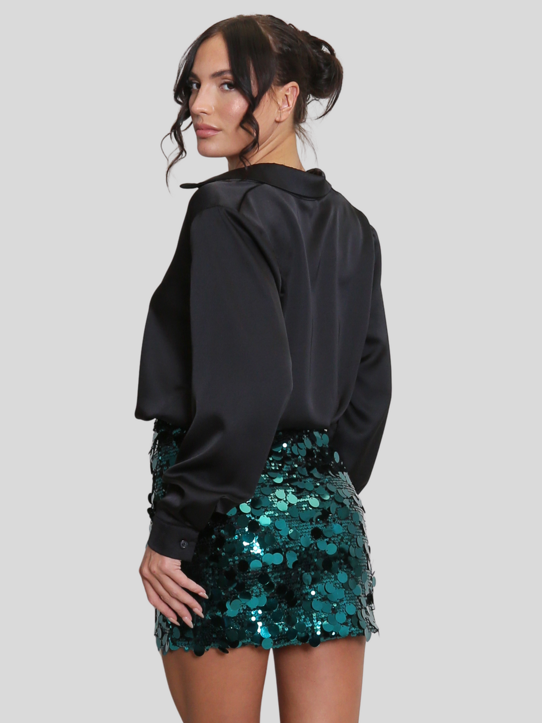 Model wears a luxe disc sequin mini skirt in green and a black shirt. The green  sequin mini skirt,  has a zip closure fastening. The model has tanned skin and black hair,  she stands with her back to the camera, the back of the skirt can be seen. 
