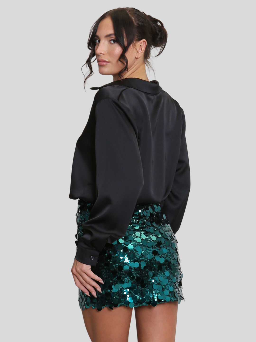 Model wears a luxe disc sequin mini skirt in green and a black shirt. The green  sequin mini skirt,  has a zip closure fastening. The model has tanned skin and black hair,  she stands with her back to the camera, the back of the skirt can be seen. 