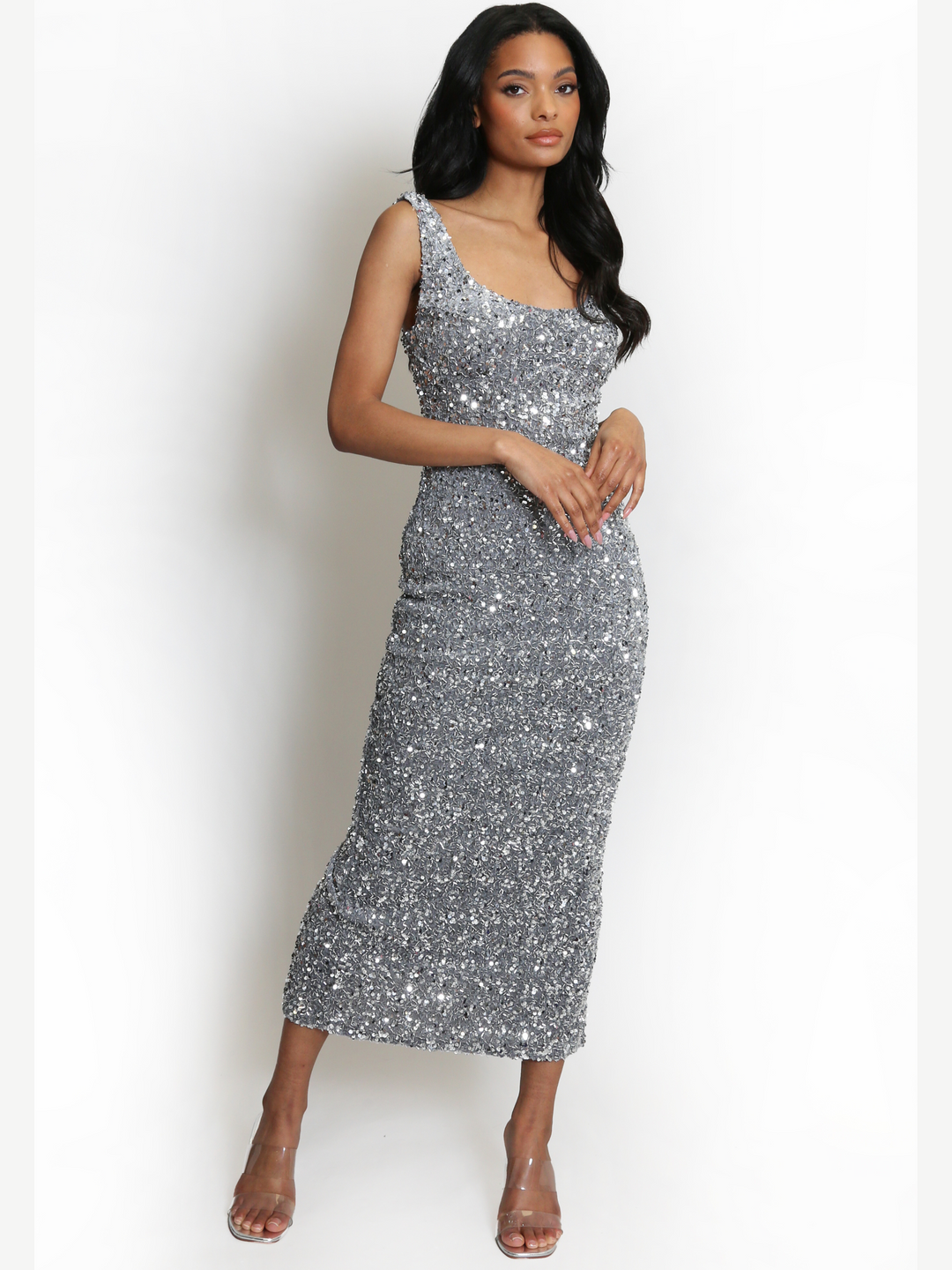 Full length shot. Model weas a  deluxe silver sequin midi dress, with a square neckline and a sleeveless cut. The model has brown skin and long black hair. The model faces the camera, the front of the sequin dress can be seen, she wears a clear perspex heel heel places her hands in front of her torso. 