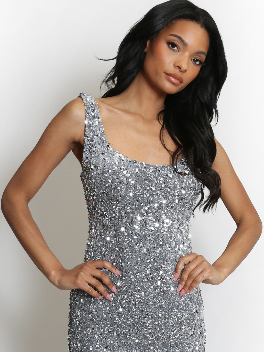 Mid length shot. Model weas a  deluxe silver sequin midi dress, with a square neckline and a sleeveless cut. The model has brown skin and long black hair. The model faces the camera, the front of the sequin dress can be seen, the model places her hands on her hips. 