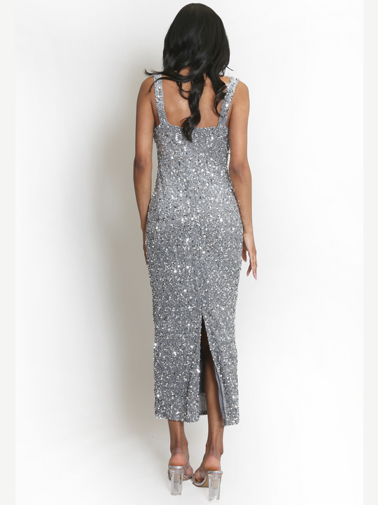 ength shot. Model weas a  deluxe silver sequin midi dress, with a square neckline and a sleeveless cut. The model has brown skin and long black hair. The model has her back to the camera, the back of the  sequin dress can be seen, including the slit at the bottom of the dress. 