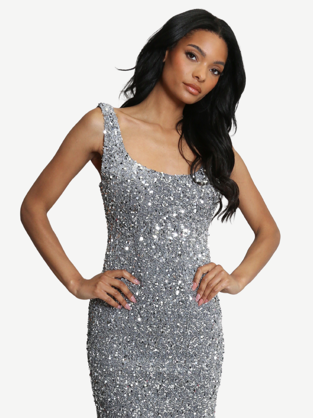 The moodel stands before a clear background. The model wears a deluxe silver sequin midi dress with a square neckline and zip back fastening. Model has brown skin and long black hair. The model stands with both hands on her hips, she tilts her head to one side and looks directly into the camera. 