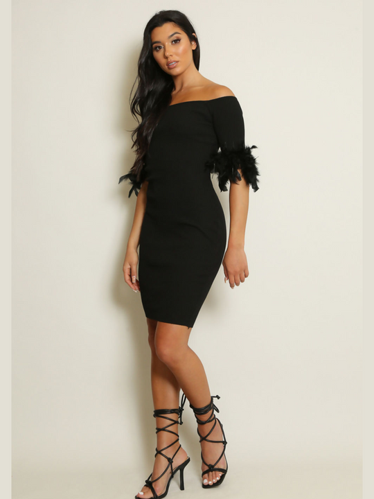 Full length shot. Woman wearing a black off-shoulder dress with feather sleeves against a plain background. The dress is a black ribbed midi dress, the model has tanned skin and dark brown hair, she stands to the side and turns her head to look into the camera. 