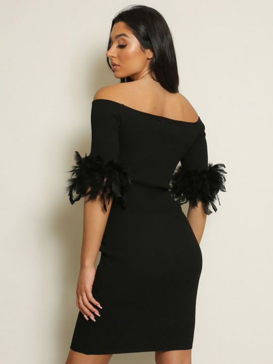 Woman wearing a black off-shoulder dress with feather sleeves against a plain background. The dress is a black ribbed midi dress, the model has tanned skin, she stands with her back to the camera, the back of the ribbed dress can be seen.