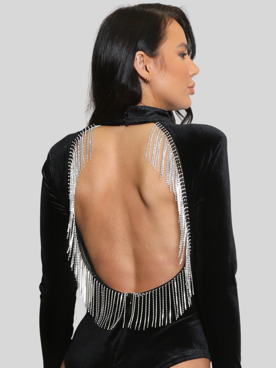 Female model wears black velvet bodysuit with an open back which has diamante tassels. The model stands in front of a plain background, the model has tanned skin and long black hair, she looks to the side. 