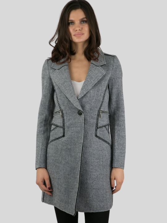 Female model stands in front of a plain background. The model wears a grey woolen coat with single front button fastening and grey tweed trim. The model has tanned skin and shoulder length hair, she faces the camera and places boh hands by her sides. The front of the coat can be seen, including teh single button fastening and exposed woven trim design and woolen lapel.
