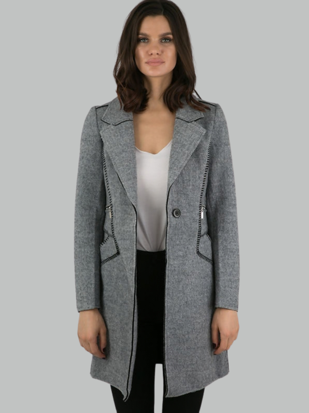 Female model stands in front of a plain background. The model wears a grey woolen coat with single front button fastening and grey tweed trim. The model has tanned skin and shoulder length hair, she faces the camera with the front of the grey woolen coat unbuttoned.  The single  grey woolen lapel can be seen. The model wears a plain white t-shirt and black trousers. 