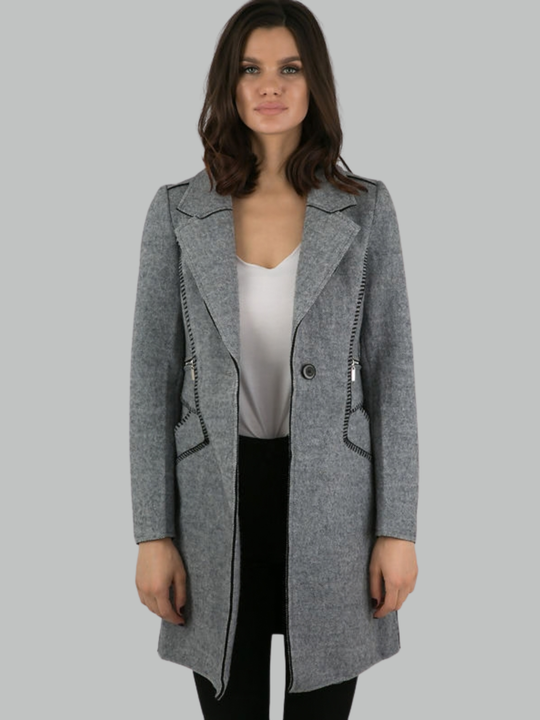 Female model stands in front of a plain background. The model wears a grey woolen coat with single front button fastening and grey tweed trim. The model has tanned skin and shoulder length hair, she faces the camera with the front of the grey woolen coat unbuttoned.  The single  grey woolen lapel can be seen. The model wears a plain white t-shirt and black trousers. 