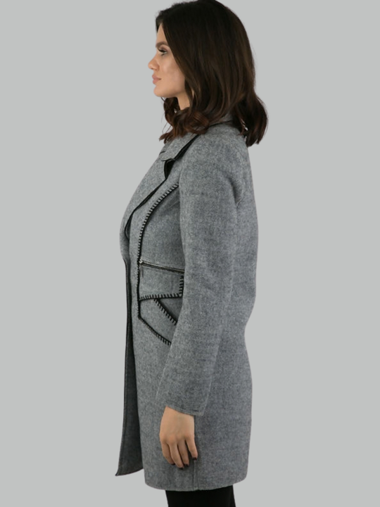 Female model stands in front of a plain background. The model wears a grey woolen coat with single front button fastening and grey tweed trim. The model has tanned skin and shoulder length hair, she stands to the side. The side of the grey coat can be seen, including the long sleeved coat and exposed kintted trim and exposed zip fastening. 