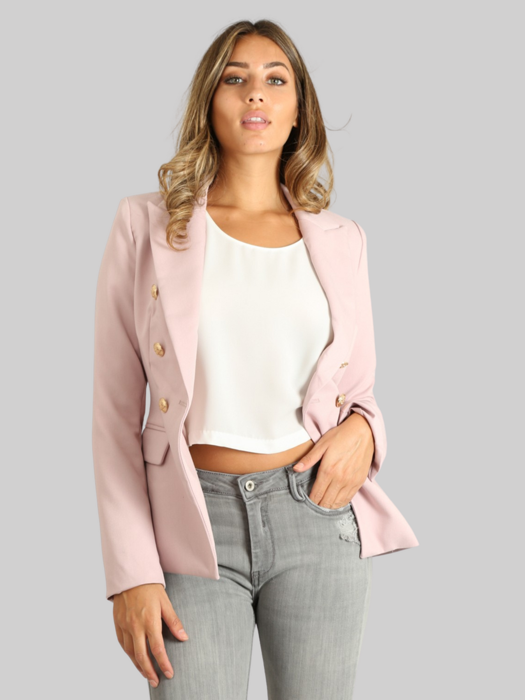 Female model wears a mauve double breatsted blazer with gold military buttons at the front of the blazer and the cuffs. The model has tanned skin and light brown hair, she faces the camera and tilts her head slightly up. The model wears the double breasted blazer open, placing one hand in her jean pocket and the other by her side. 