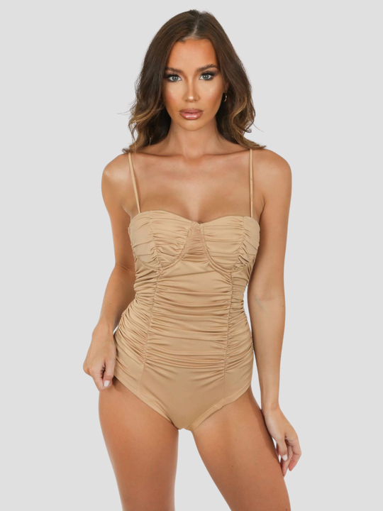 Female model stands before a plain background. The model wears a nude bodysuit with a sweetheat neckline and cup detailing and adjustable spagehtti straps. The model has tanned skin and brown hair, she faces the camera and places one hand on her hip and the other by her side, the ruched detailing can be seen. 