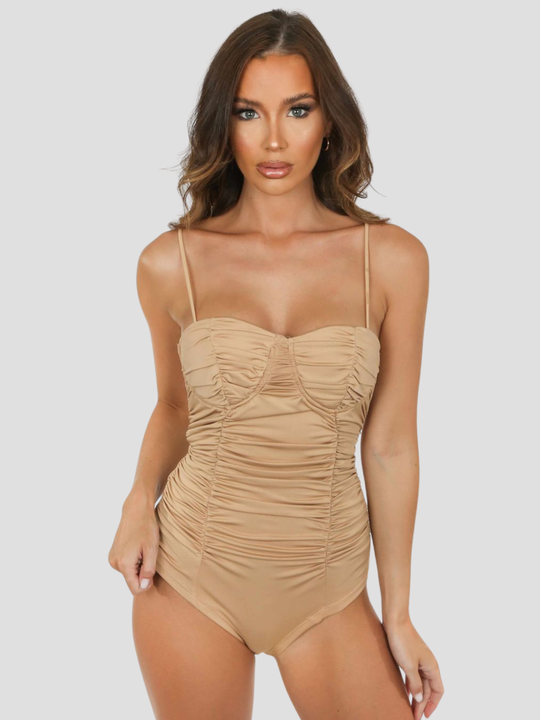 Female model stands before a plain background. The model wears a nude bodysuit with a sweetheat neckline and cup detailing and adjustable spagehtti straps. The model has tanned skin and brown hair, she faces the camera and places one hand on her hip and the other by her side. 