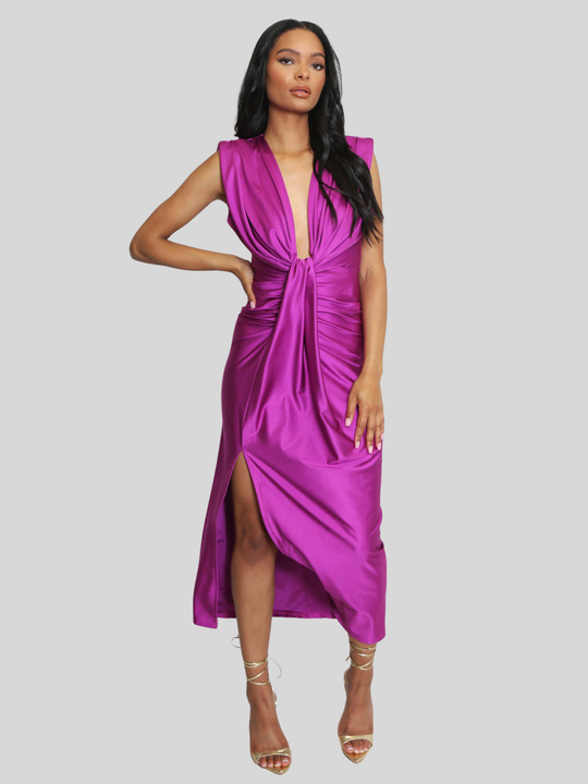 Model stands in front of a plain background, she wras a drape front fuschia occasion midi dress, which is sleeveless and has a side thigh split design. The dress has a ruched front. The model has light brown skin and long black hair, the model places one hand on her hip and the other on her thigh, she wears strappy high heeled shoes.