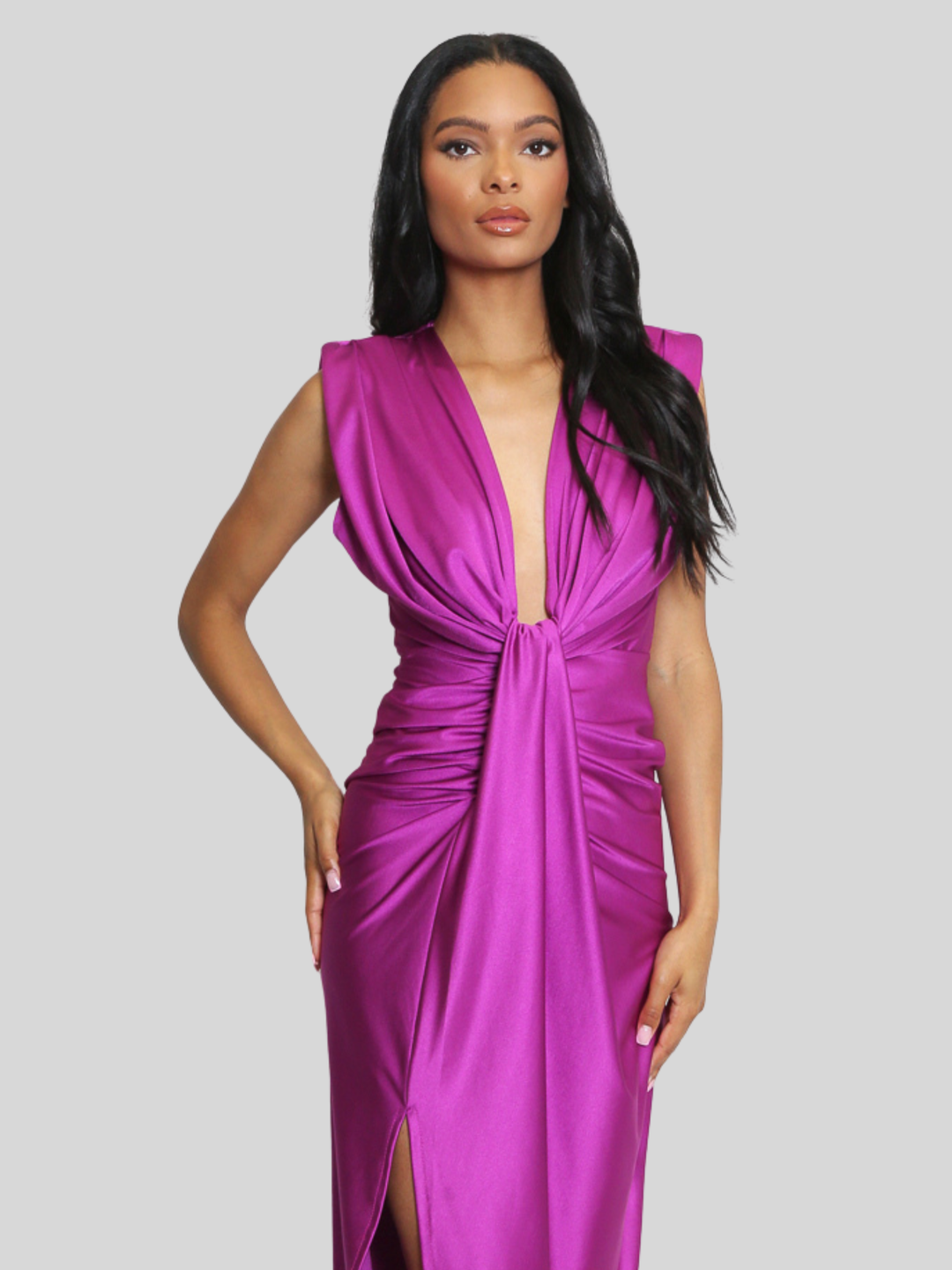Model stands in front of a plain background, she wras a drape front fuschia occasion midi dress, which is sleeveless and has a side thigh split design. The dress has a ruched front. The model has light brown skin and long black hair, she places one hand on her hop and the other by her side, the drape front design can be seen. 