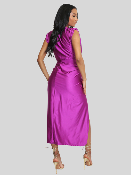 Full length shot. Model stands in front of a plain background, she wras a drape front fuschia occasion midi dress, which is sleeveless and has a side thigh split design. The dress has a ruched front. The model has light brown skin and long black hair, she stands with her back to the camera, the back of the dress and her strappy heels can be seen. 