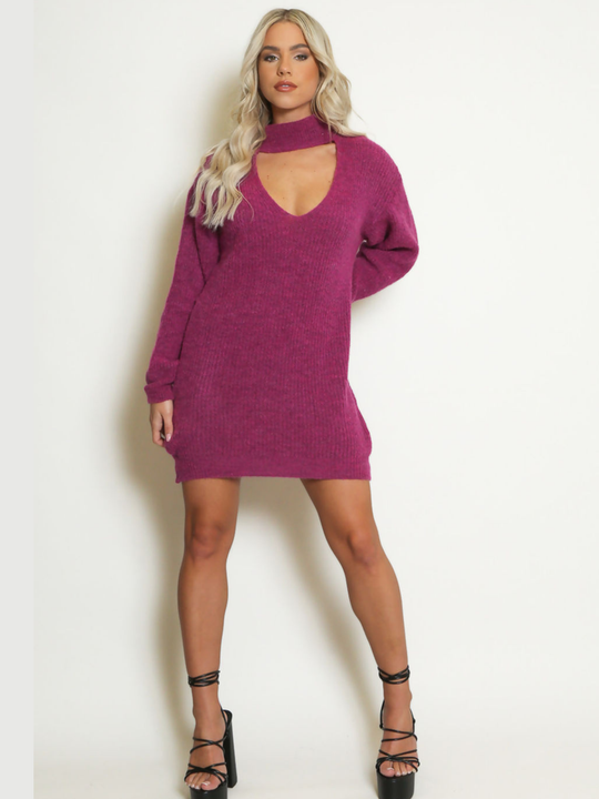 Full length shot. emale model stands in front of a plain background. The model wears a purple knitted long sleeved mini jumper dress. The jumper dress has a high neck and cut out front. The model has tanned skin and wavy blonde hair, she faces the camera and places one hand behind her back and the other by her side. The model wears black lace up platform heels. 