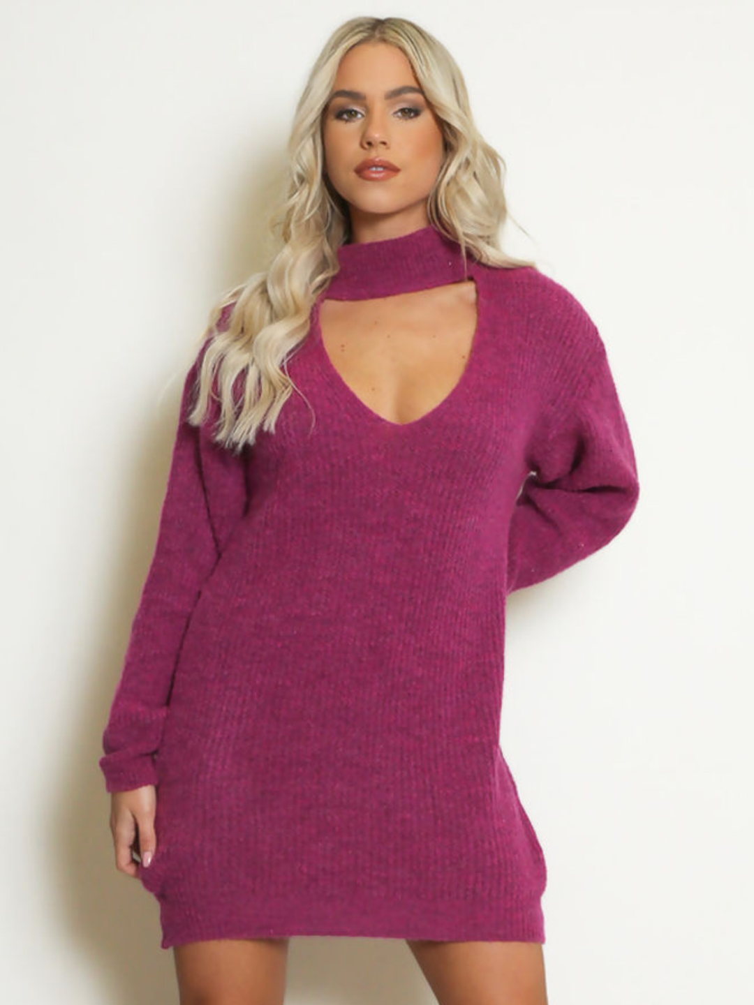 Female model stands in front of a plain background. The model wears a purple knitted long sleeved mini jumper dress. The jumper dress has a high neck and cut out front. The model has tanned skin and wavy blonde hair, she faces the camera and places one hand behind her back and the other by her side. 