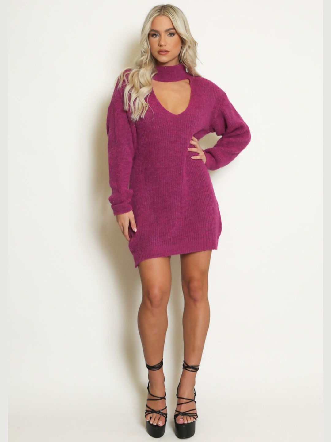 Full length shot. Female model stands in front of a plain background. The model wears a purple knitted long sleeved mini jumper dress. The jumper dress has a high neck and cut out front. The model has tanned skin and wavy blonde hair, she faces the camera and places on hand on her hip and the other by her side. The model wears black lace up platform heels. 