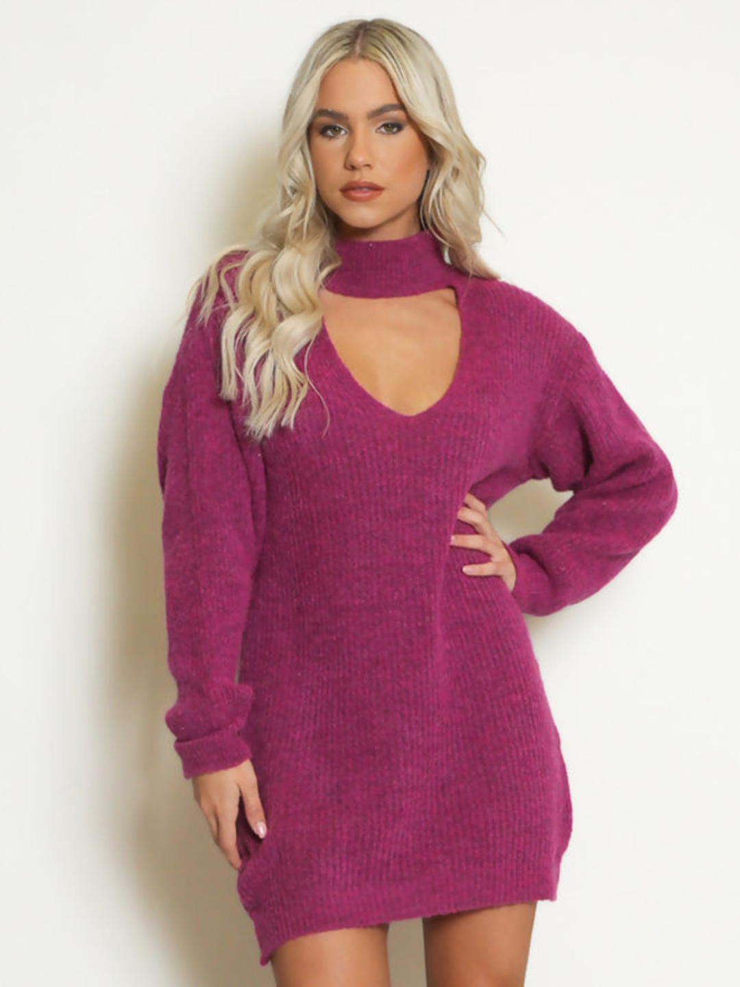 Female model stands in front of a plain background. The model wears a purple knitted long sleeved mini jumper dress. The jumper dress has a high neck and cut out front. The model has tanned skin and wavy blonde hair, she faces the camera and places on hand on her hip and the other by her side. 