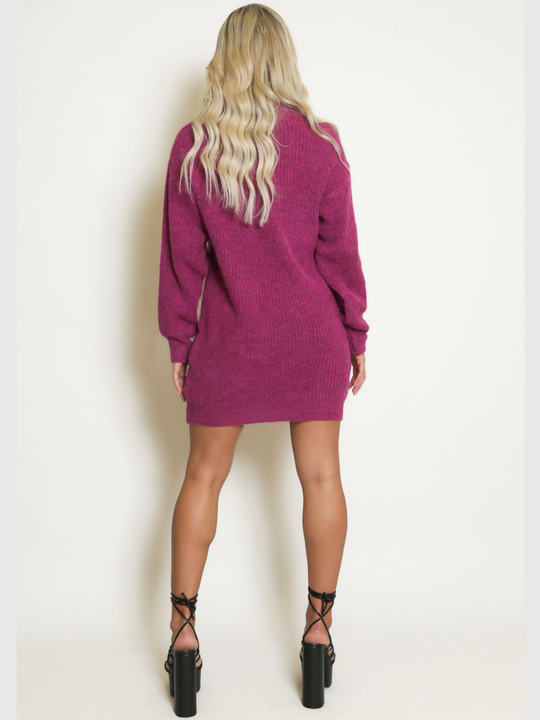 Full length shot. Female model stands in front of a plain background. The model wears a purple knitted long sleeved mini jumper dress. The jumper dress has a high neck and cut out front. The model has tanned skin and wavy blonde hair, she stands with her back to the camera, the ribbed knitted design of the back of the jumper dress can be seen. The model wears black lace up high heels with a chunky heel design. 