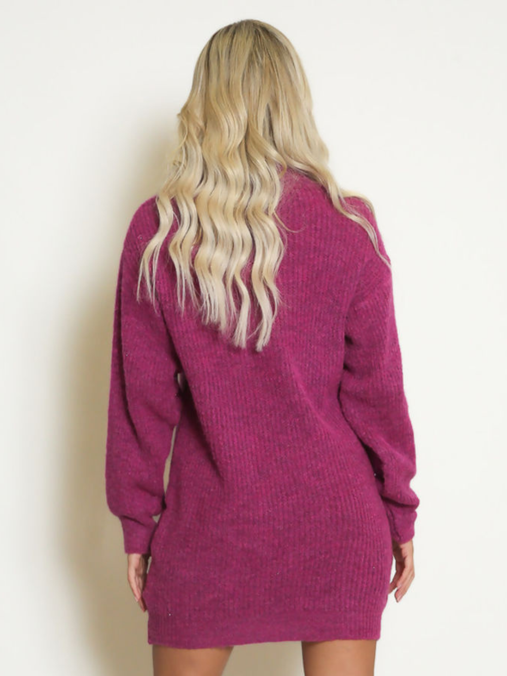 Female model stands in front of a plain background. The model wears a purple knitted long sleeved mini jumper dress. The jumper dress has a high neck and cut out front. The model has tanned skin and wavy blonde hair, she stands with her back to the camera, the ribbed knitted design of the back of the jumper dress can be seen. 
