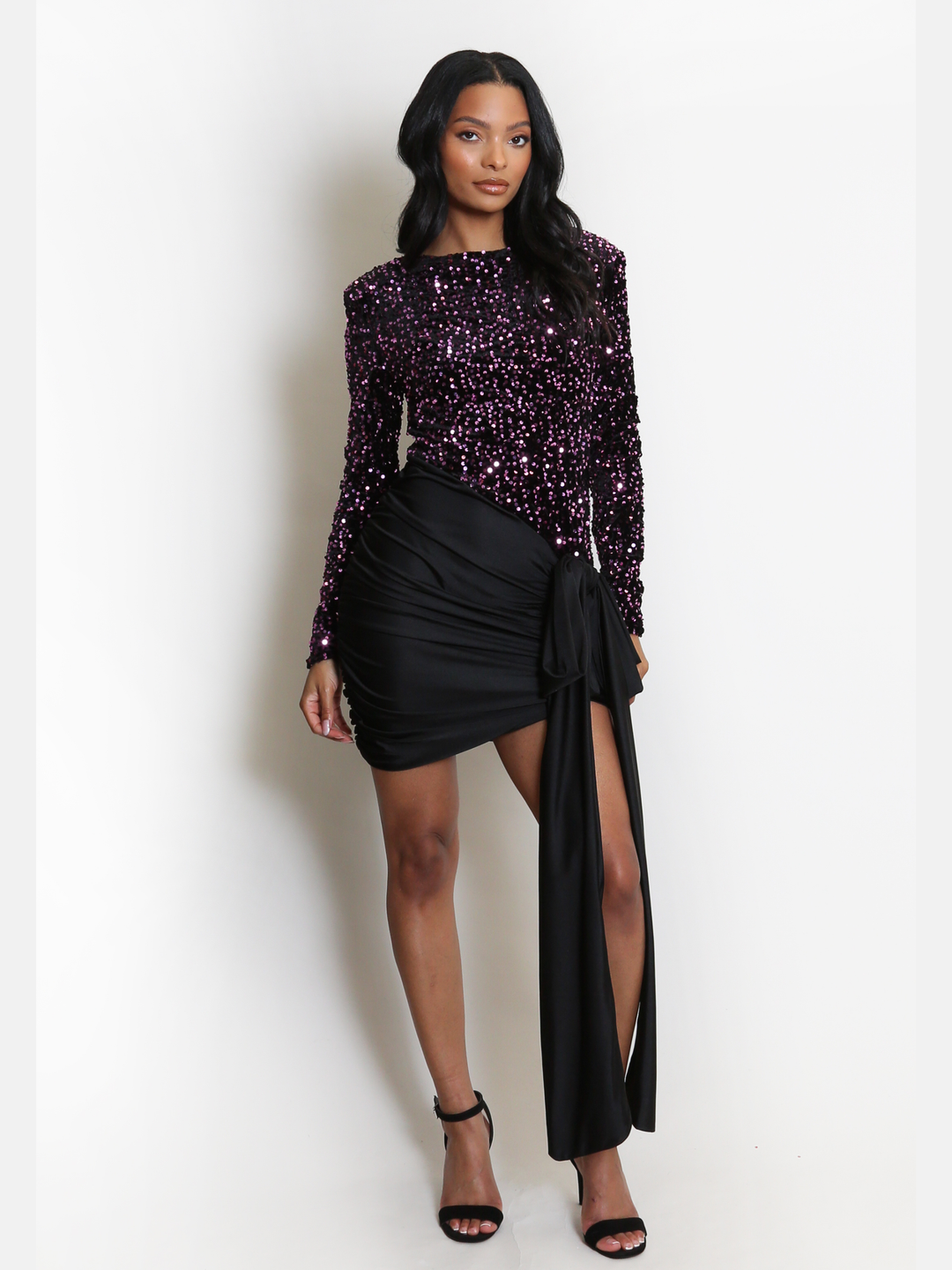 Full length shot. Model stands in front of a plain background. The model wears an occasion dress that has a purple sequinned long sleeve bodice and a black ruched satin mini fit,  with drape detailing. The model has light brown skin and long black hair, she places one eg sightly in front of the other and both hands by her sides, the satin drape detailing can be seen. 
