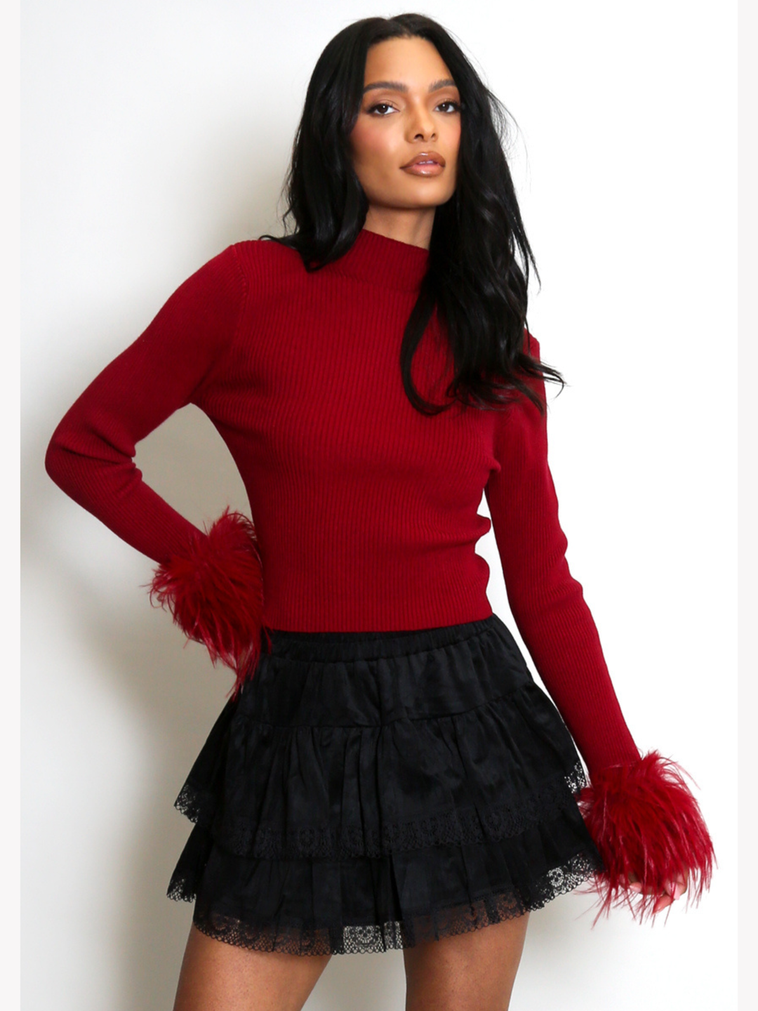 Model stands in front of a plain cream background. The model wears a red fine knit ribbed jumper with red faux fur cuffs and a black lace ruffled skirt. The model has light brown skin and long black hair, she faces the camera, placing one hand on her hip and the other by her side. The faux fur cuffs can be seen. 