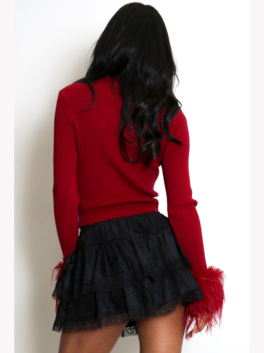 Model stands in front of a plain cream background. The model wears a red fine knit ribbed jumper with red faux fur  cuffs and a black lace ruffled skirt.  The model has light brown skin and long black hair, she stands with her back to the camera, the back of the fine knit ribbed jumper can be seen. 