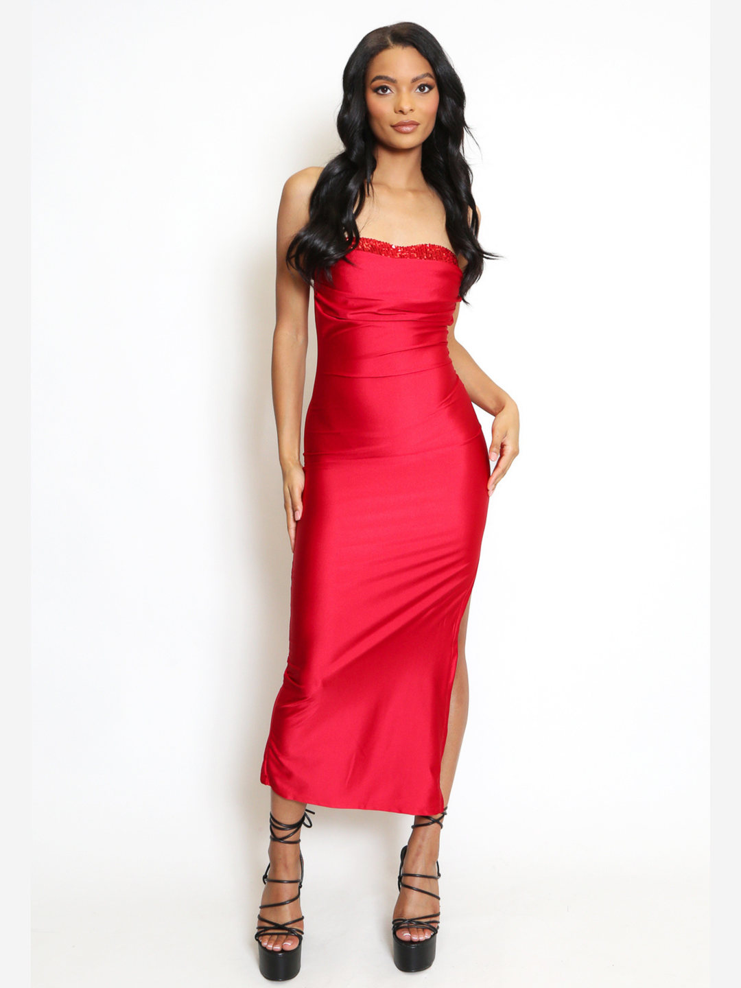 Full shot. Female model stands in front of a plain background, she wears a red midi dress that has a ruched cup design and red bandeau sequin front insert. The midi dress also has a side split. The model has light brown skin and long black hair, she faces the camera, placing one hand by her side and the other on her hip. The model wears black plaform strappy heels. 