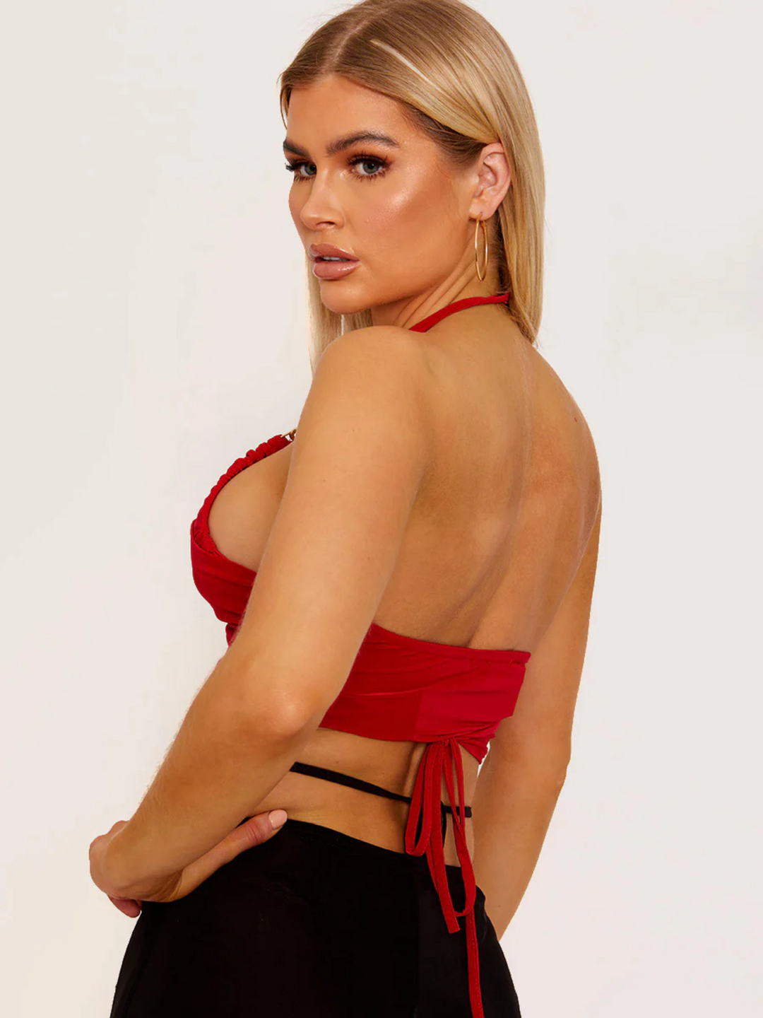 This image showcases a striking red wrap-style crop top, featuring an alluring halter neck and sophisticated gold chain detailing on the straps. The crop top's design is both elegant and bold, with a snug fit that accentuates the silhouette, making it a standout piece for evening wear or special occasions. Model has tanned skin and blonde hair, she stands to the side, the back and side if the halter neck can be seen, including the the tie fastening. Model looksaway from the camera.