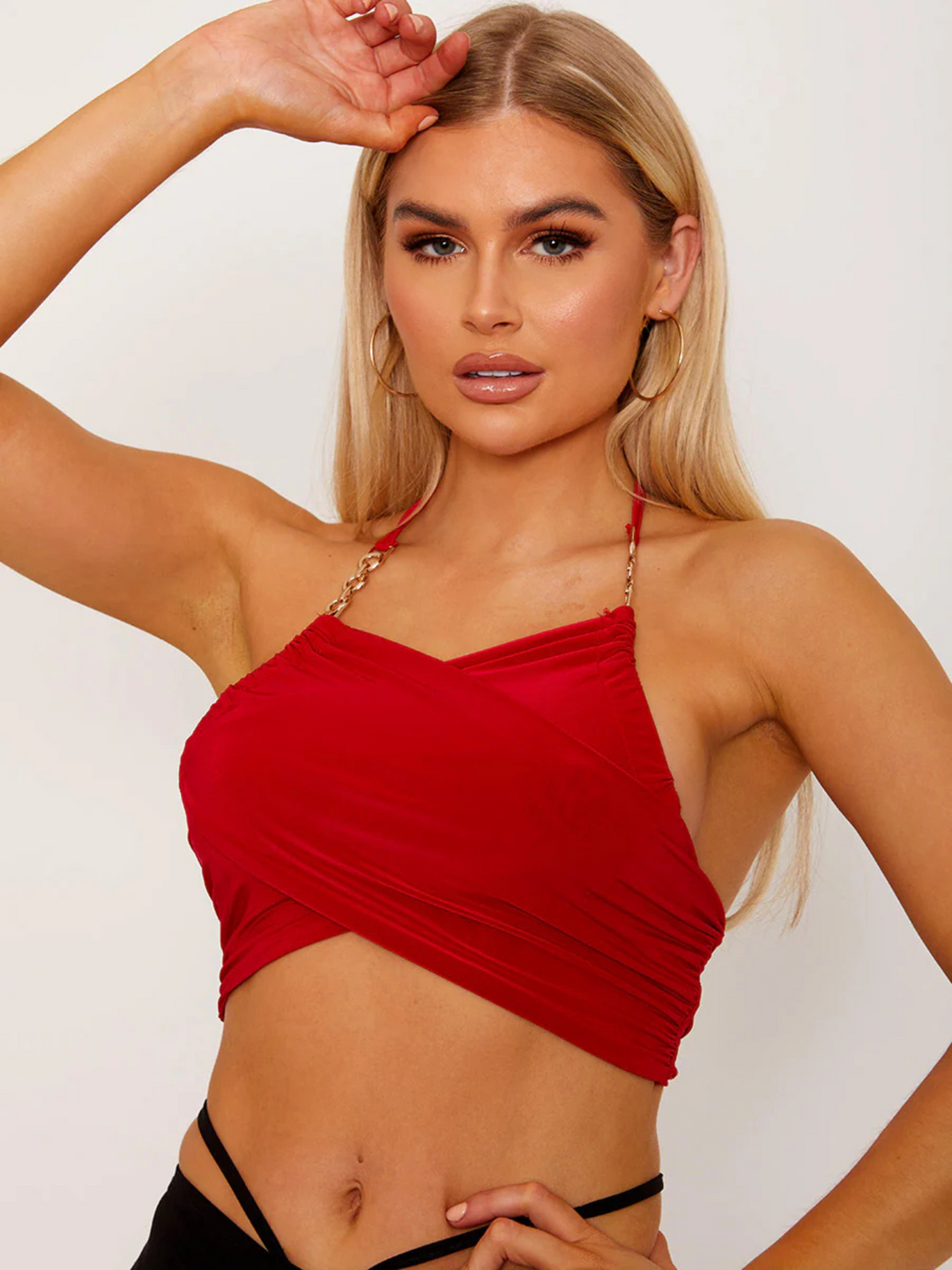 This image showcases a striking red wrap-style crop top, featuring an alluring halter neck and sophisticated gold chain detailing on the straps. The crop top's design is both elegant and bold, with a snug fit that accentuates the silhouette, making it a standout piece for evening wear or special occasions. Model has tanned skin and blonde hair, she places one hand on her hip and the other on her head.
