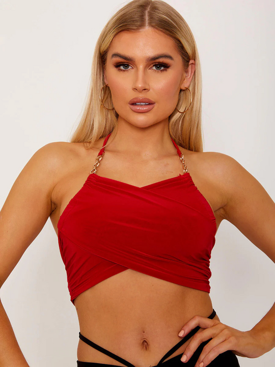 This image showcases a striking red wrap-style crop top, featuring an alluring halter neck and sophisticated gold chain detailing on the straps. The crop top's design is both elegant and bold, with a snug fit that accentuates the silhouette, making it a standout piece for evening wear or special occasions. Model has tanned skin and blonde hair, she faces forward, looking directly into the camera, placing her hand on her hips.
