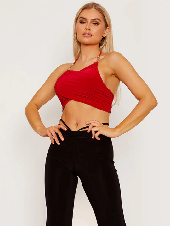 This image showcases a striking red wrap-style crop top, featuring an alluring halter neck and sophisticated gold chain detailing on the straps. The crop top's design is both elegant and bold, with a snug fit that accentuates the silhouette, making it a standout piece for evening wear or special occasions. Model has tanned skin and blonde hair, she faces the camera, places his hands on his hips. Model looks directly into the camera.