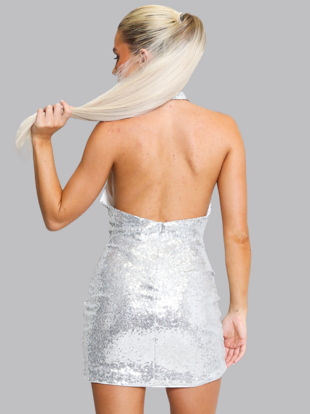 Female model wears a silver sequin cowl neck halter mini dress. She stands in front of a plain grey background. The model has  tanned skin and hair that she pulls back into a ponytail. The model stands with her back to the camera, the halter neck back of the dress can be seen. The model holds her blinde ponytail in one hand and places her other arm by her side. 