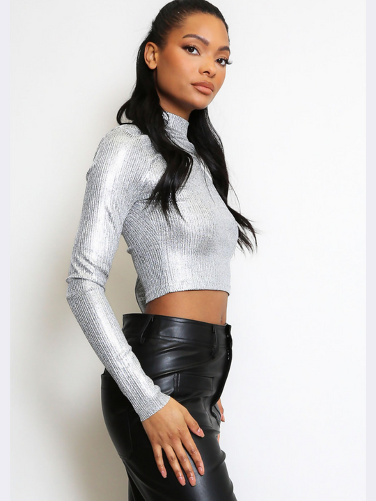 This image features a striking silver metallic crop top, characterized by its high neck and long sleeves. The crop top's shiny, reflective surface captures light, creating a vibrant and eye-catching effect. The model has brown skin and long brown hair, sher stands to the side, and turns her head to look into the camera,  the side of the crop top can be seen.