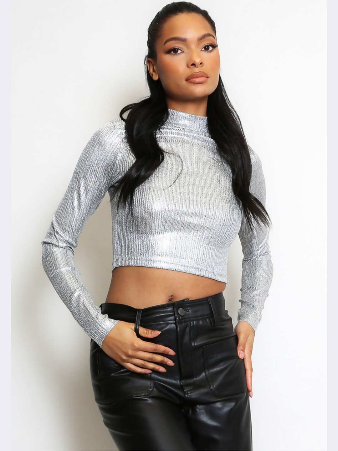 This image features a striking silver metallic crop top, characterized by its high neck and long sleeves. The crop top's shiny, reflective surface captures light, creating a vibrant and eye-catching effect. The model has brown skin and long brown hair, she faces the camera placing one hand by her side and the other on her thigh. 