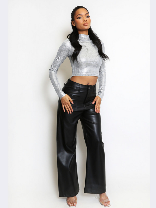 Full length shot. The female model wears  a striking silver metallic crop top, characterized by its high neck and long sleeves. The crop top's shiny, reflective surface captures light, creating a vibrant and eye-catching effect. The model has brown skin and long brown hair, she faces the camera and places both hands on her thighs, the front of her  black pu trousers can be seen. 